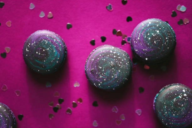 A close-up of colorful, galaxy-themed macarons scattered on a vibrant pink background with small heart-shaped glitter