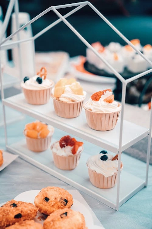 A white dessert display stand with three tiers showcasing cupcakes topped with whipped cream and fresh fruit